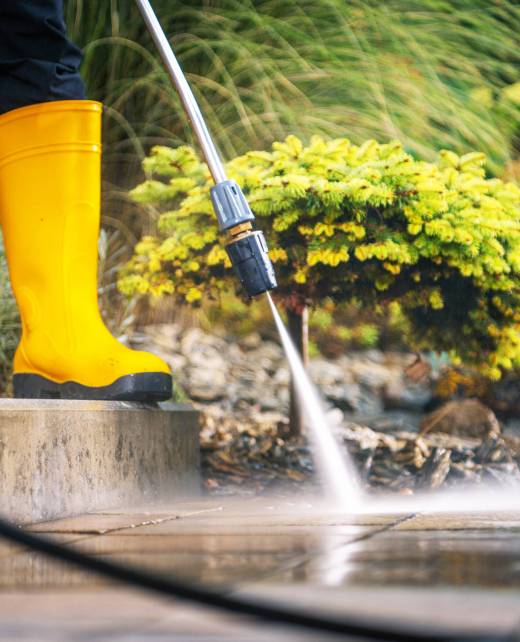 A person is using a pressure washer to clean a patio surface while standing in a beautifully landscaped garden. The vibrant greenery and flowers create an inviting atmosphere.