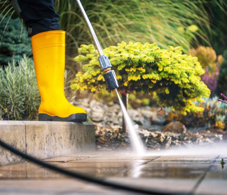 A person is using a pressure washer to clean a patio surface while standing in a beautifully landscaped garden. The vibrant greenery and flowers create an inviting atmosphere.
