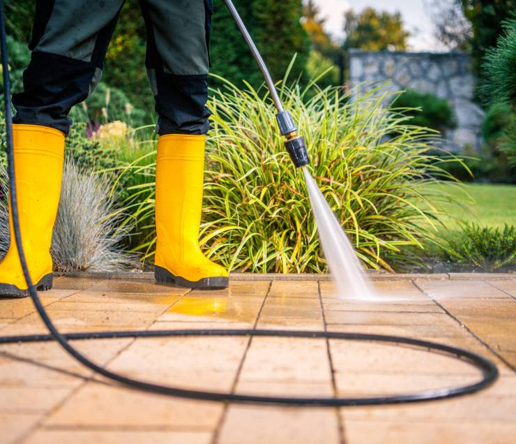 A person in yellow boots operates a pressure washer to clean a patio, surrounded by colorful plants in a well-maintained garden during daytime.